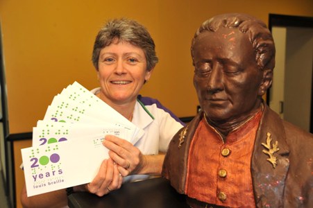 woman holding commemorative braille envelopes and standing beside a statue of Louis Braille