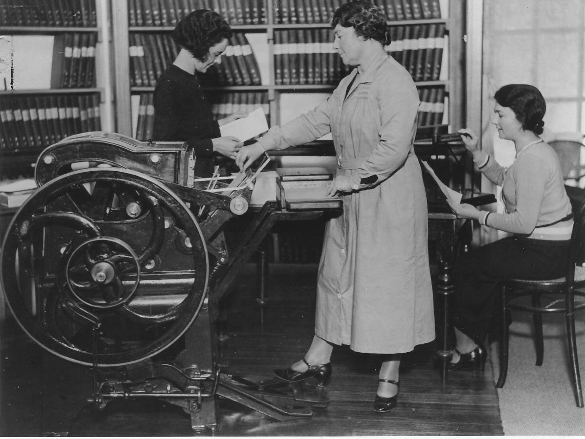 Miss Minnie Crabb operating an cast iron Braille printing machine using her foot. Library shelves are featured in the background.