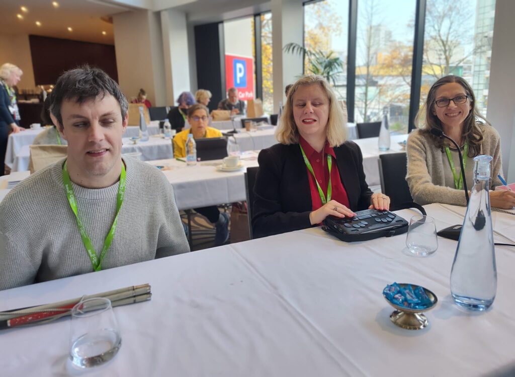 Scott Erichsen, Jordie Howell and Jodie Doolan at the 2024 ICEB General Assembly. They are smiling and seated at a table with microphone stand.